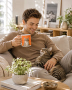 A man in a brown sweater relaxes on a beige sofa with a tabby cat on his lap, holding a white ceramic mug with an orange graphic and a cat illustration. The scene is set in a bright, modern living room with plants and wooden furniture.