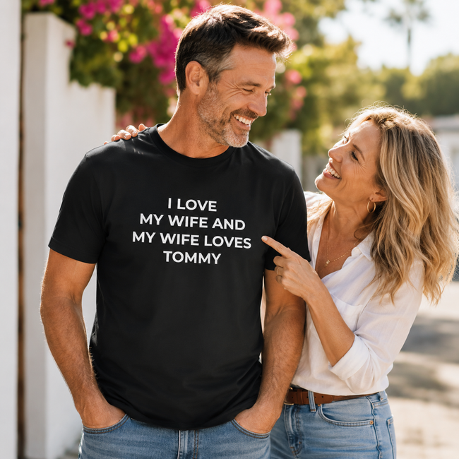 A man wearing a black graphic t-shirt with white text that reads 'I LOVE MY WIFE AND MY WIFE LOVES TOMMY' is being pointed at by a woman in a white blouse and jeans, set against an outdoor background with blurred pink flowers and green foliage. Hover Image