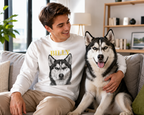 A man and a husky dog sit together on a gray couch in a bright living room. The man is wearing a white long-sleeve crewneck sweatshirt with a custom graphic of a husky dog's face and the name 'BILLY' printed in yellow above it.
